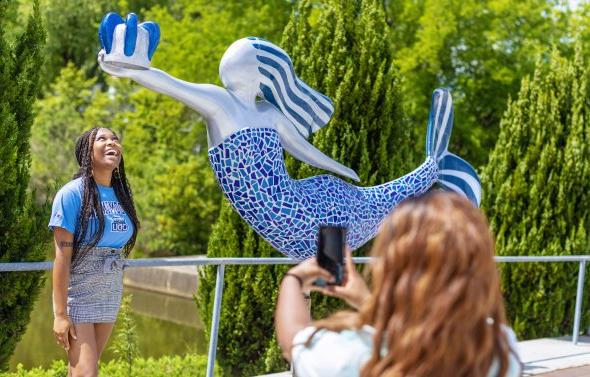 一名身穿蓝色衬衫的学生站在诺福克美人鱼雕像下，另一名学生正在给她拍照