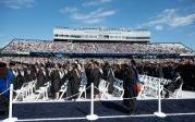 Students wearing academic robes and mortarboards stand in rows in a stadium.